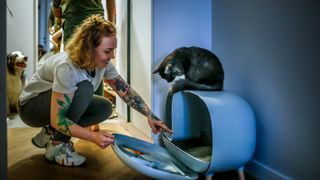 A woman opening a hooded litter box which a cat is sitting on top of and looking down into