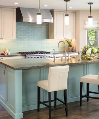 A kitchen with a teal blue splashback and kitchen island which has a black granite surface and sink, two cream bar chairs and three hanging pendant lights above it