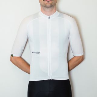 A white man in a white cycling jersey and black cycling shorts stands against a white wall