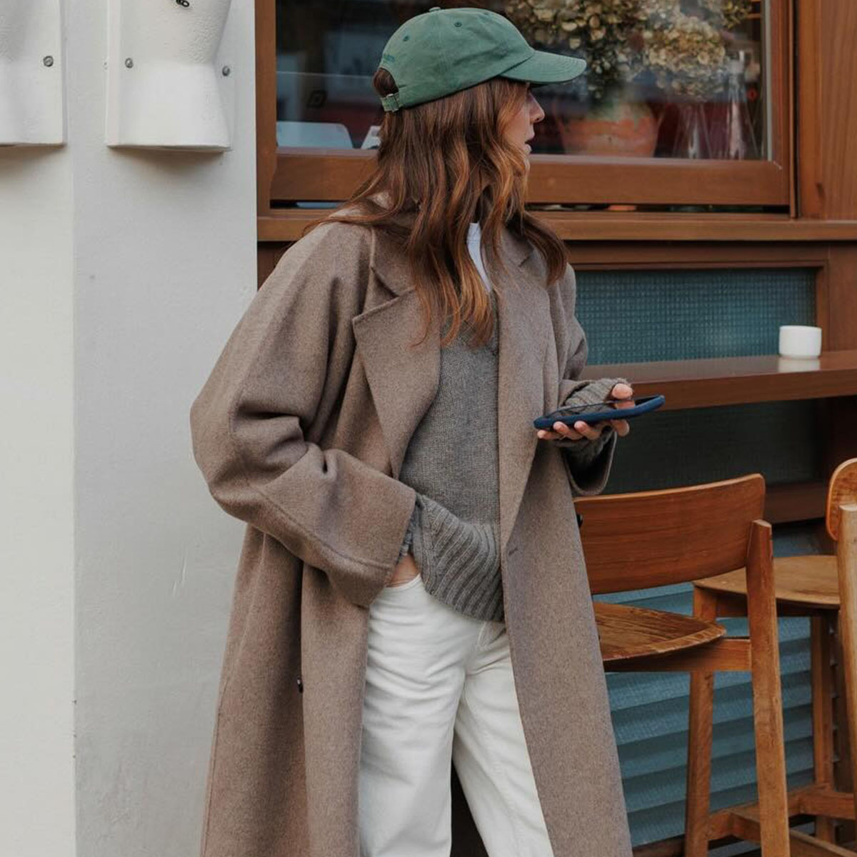 Woman in a beige coat, gray sweater, green hat, and white jeans