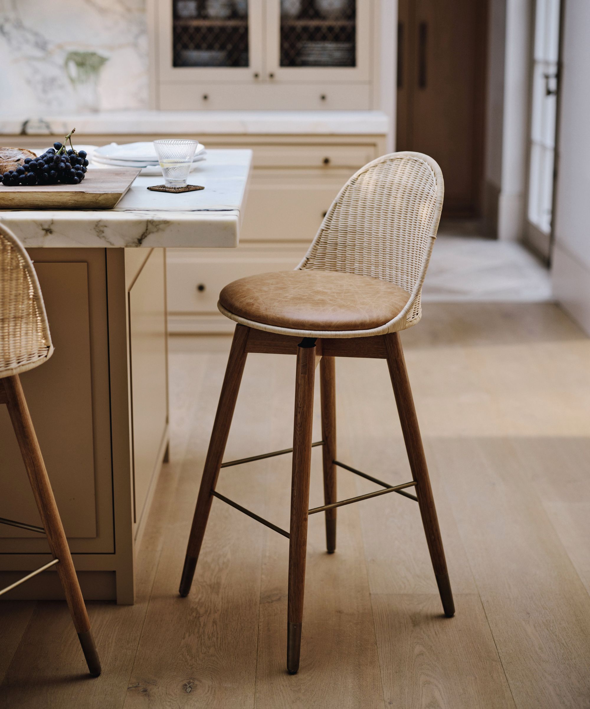 Close-up of a woven rattan bar stool with a tan leather seat beside a marble-topped kitchen island, set on pale wood flooring
