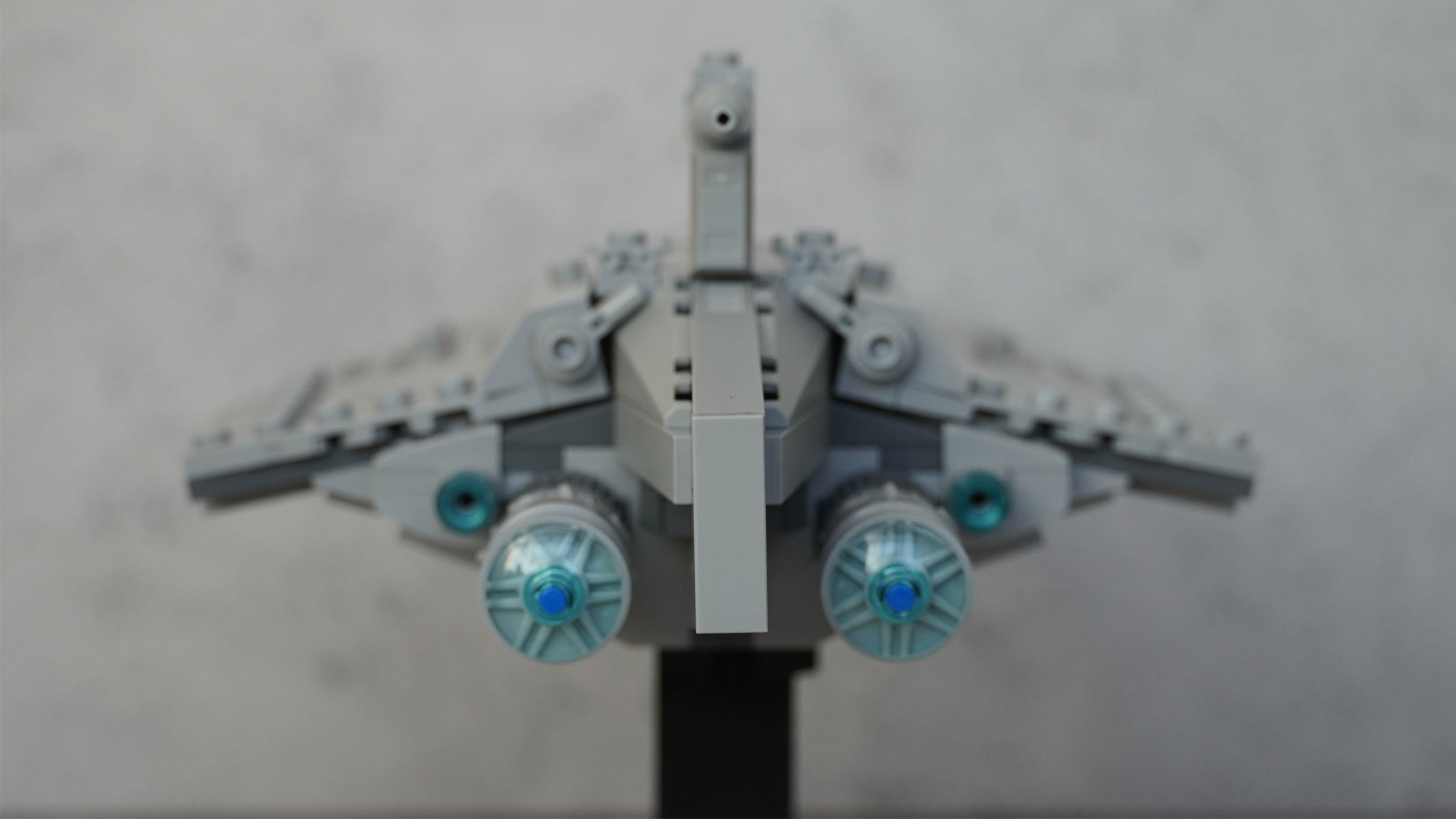 A miniature-sized spaceship made of grey bricks is on display with rear engines on display. It's placed on a black stand in front of a grey background.