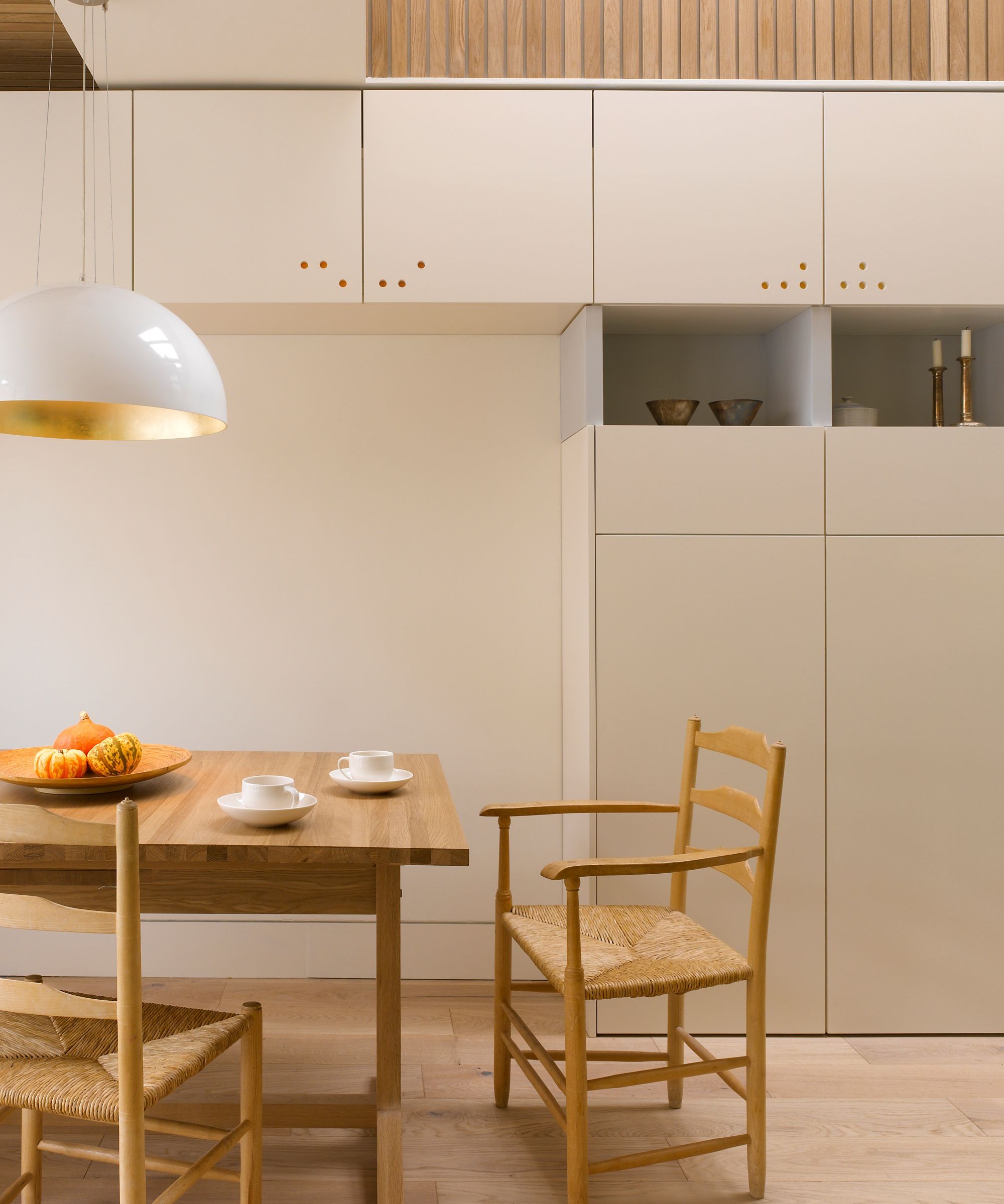 A kitchen with wood cladding on the wall, plain stone coloured cupboards and wooden chairs and tables in the foreground
