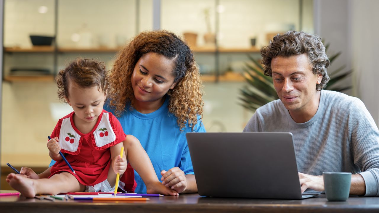 A young couple sits at their dining room table while she keeps their toddler occupied and he works on his laptop.