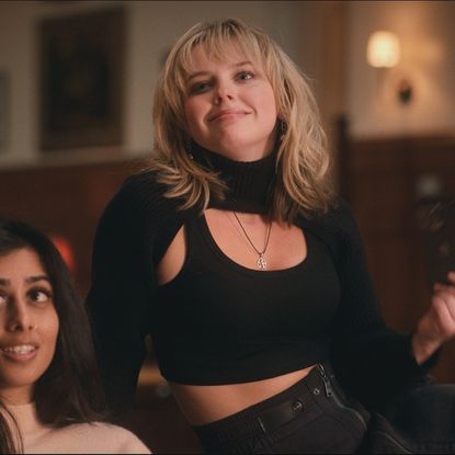 Mia Rodgers, right, sits on the leg of a chair next to a girl on her left, in 'Sex Lives of College Girls' season 3.