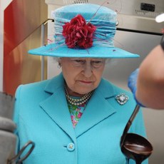 Queen Elizabeth wears a turquoise suit and matching hat and watches a chef serve something with a large metal ladle