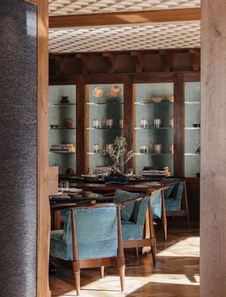 A transitional design style hotel restaurant features classic blue velvet and wood dining chairs, sun-lit wooden floors, and a wood and glass shelving unit filled with collectibles.