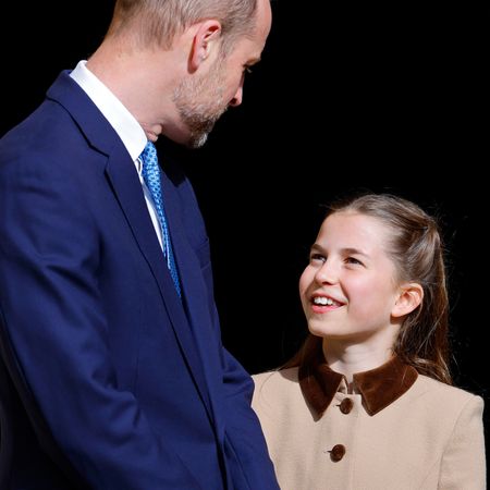 Princess Charlotte wears a two-tone brown coat dress and gazes up at dad Prince William while smiling, she has long brown hair, while her dad wears a navy suit and is balding