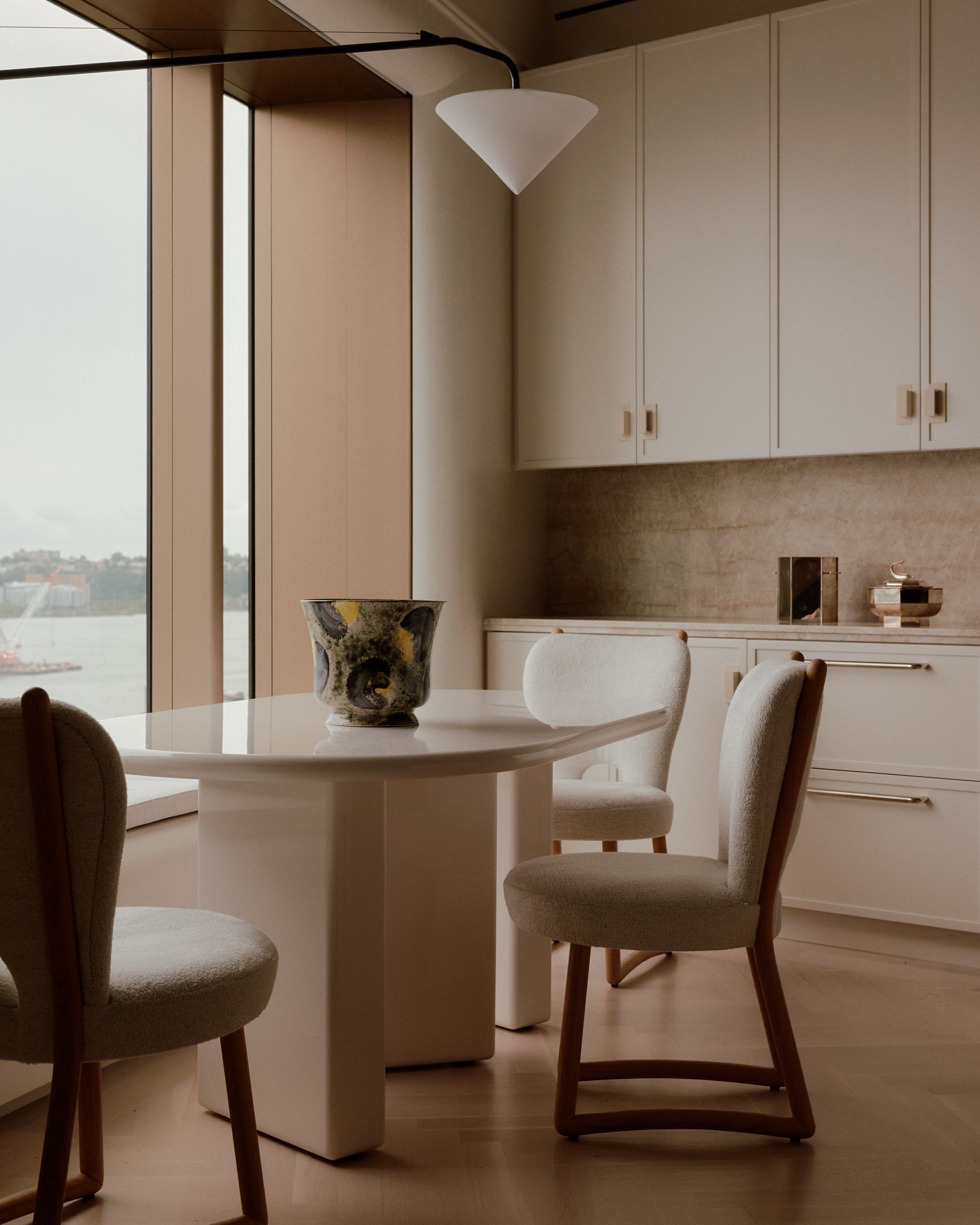 neutral kitchen with small dining nook with table and three chairs, styled with a vase. behind is the kitchen running across the back wall, with white cabinets and a neutral-colored stone backsplash