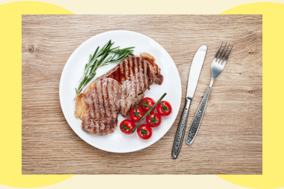 A plate of food with sirloin steak, rosemary and tomatoes on a wooden table with cutlery