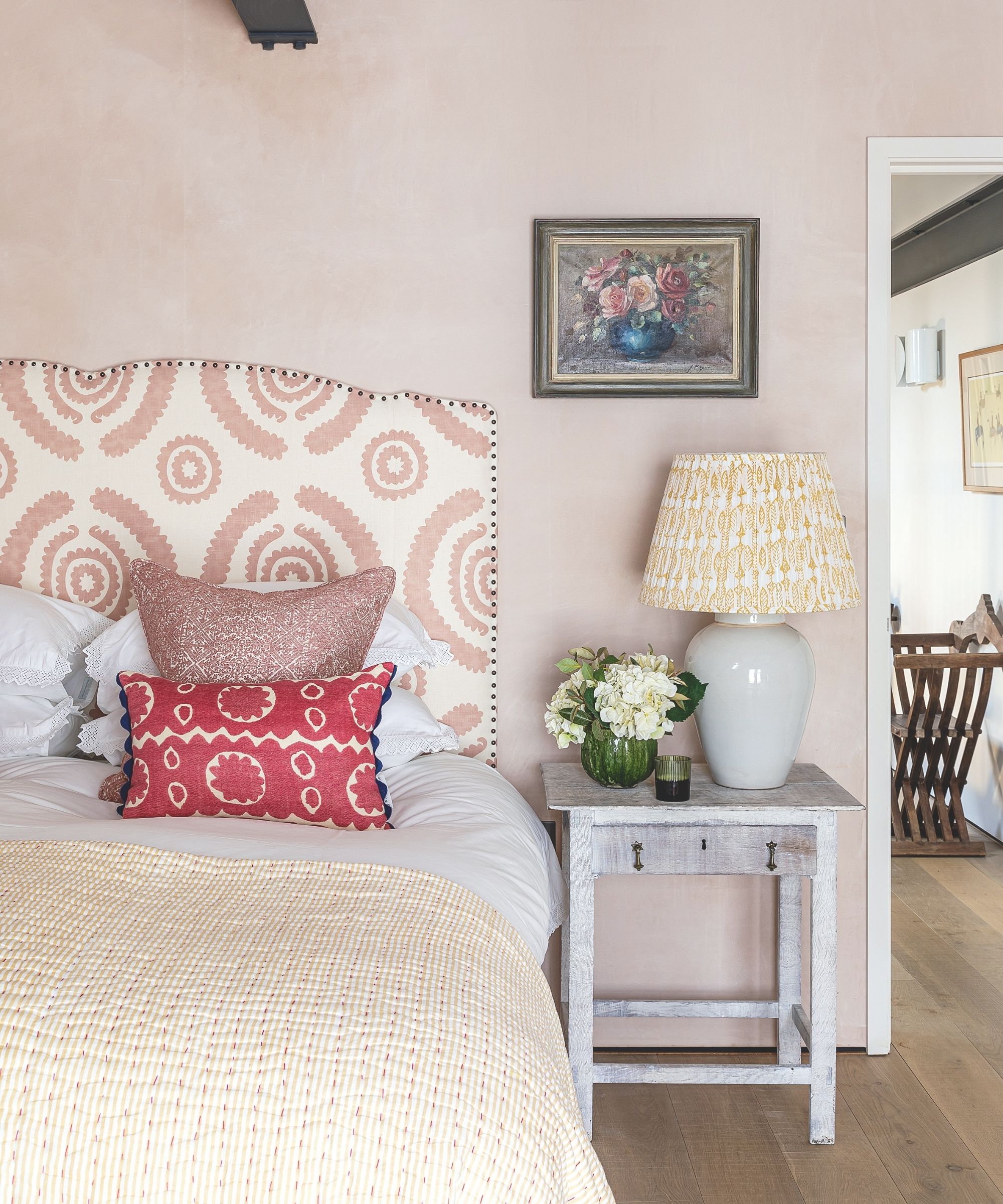 Pale pink bedroom with patterned headboard, bedside table, vase of white flowers, white ceramic lamp with white and yellow patterned shade, and a small still life artwork of flowers above.