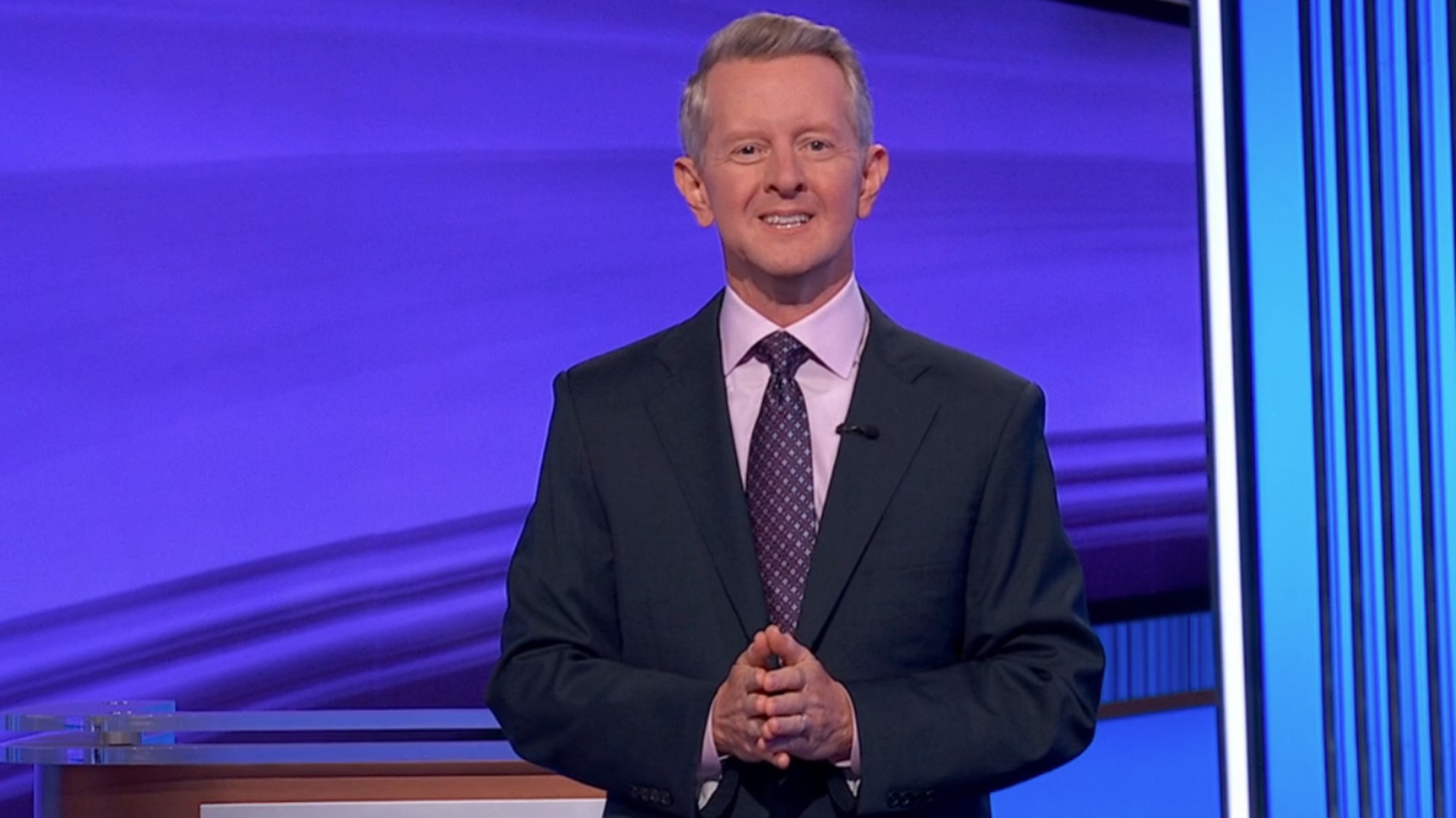 Ken Jennings at the beginning of an epsiode of Jeopardy, wearing a blue suit, pink shirt and blue and pink tie