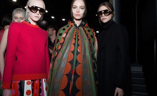 Three models, one in black, one in red and one in a black, green, orange and brown outfit with geometric designs