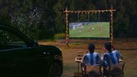 A car projecting a football match from its headlights onto a screen outside with two people sitting in chairs watching.