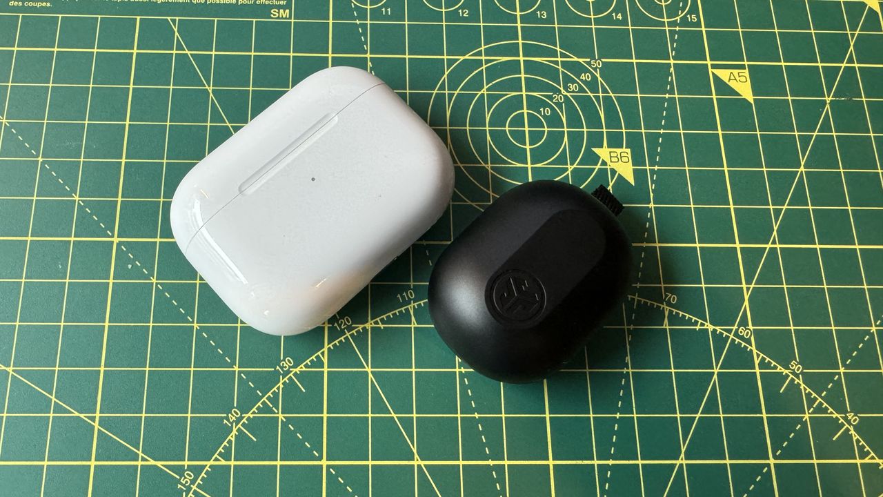 JLab JBuds Mini true wireless headphones on a book and technical mat, with hand and AirPods for comparison.