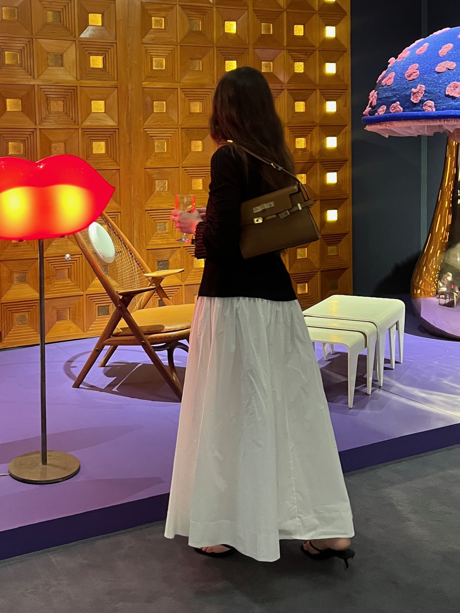 Caitlin Burke at an art exhibition wearing a black blazer and white skirt and carrying a Saint Laurent Manhattan bag in brown.
