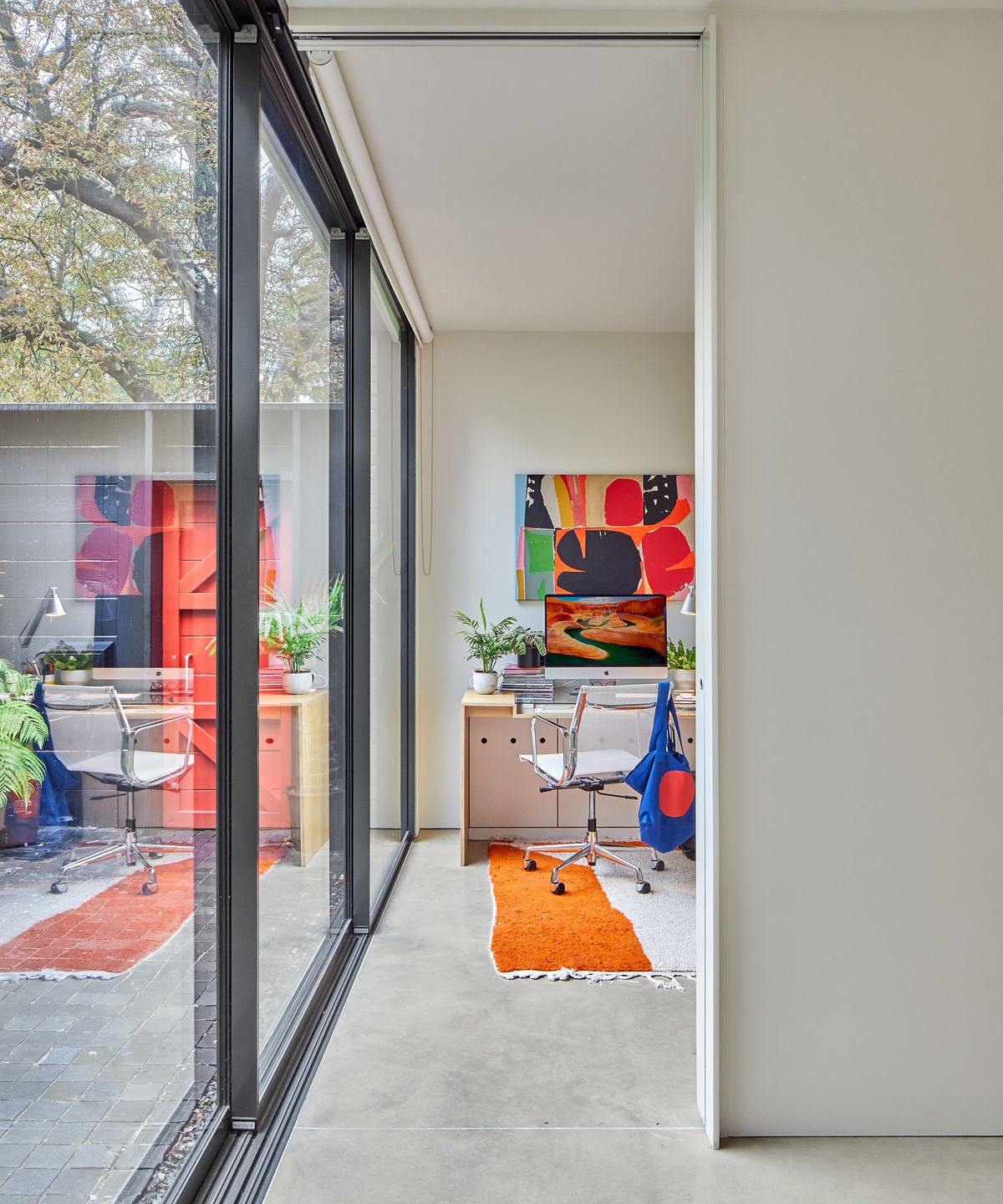 A small home office on the ground floor of a contemporary home. The entire wall to the left and side is made of floor-to-ceiling glazing. The decor of the office is light with pops of colour from art prints and a rug