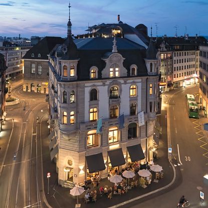 Exterior image of Hotel M&auml;rthof Basel shows a building with tables and chairs and umbrellas outside, with tramlines running past the hotel
