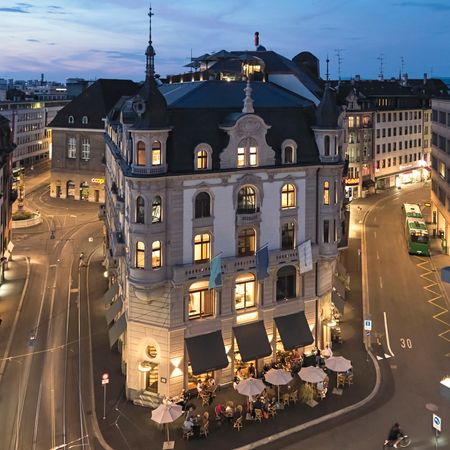 Exterior image of Hotel M&auml;rthof Basel shows a building with tables and chairs and umbrellas outside, with tramlines running past the hotel