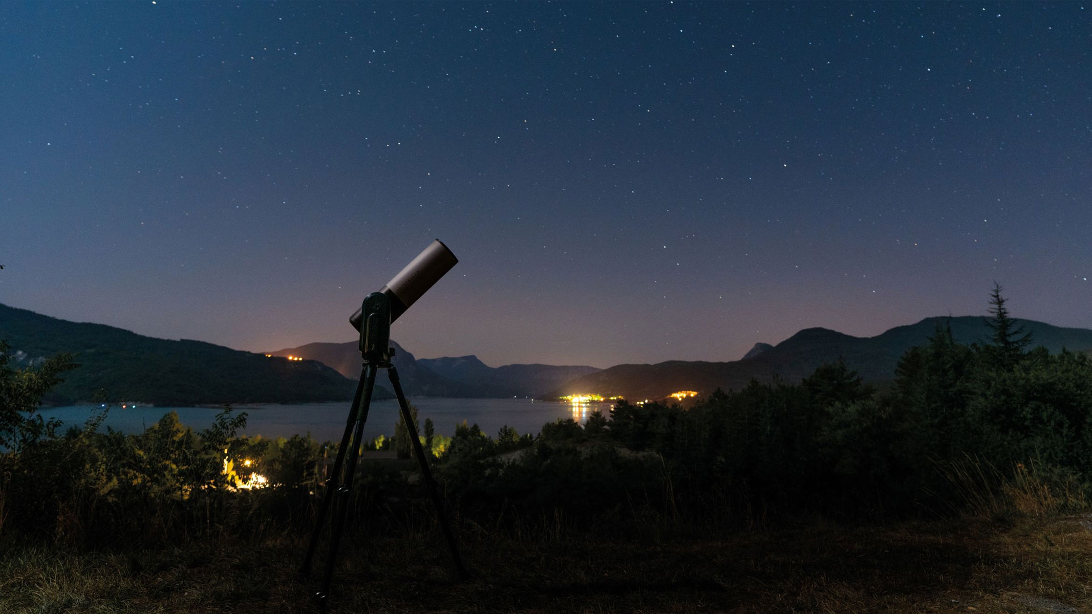 A Unistellar smart telescope with the backdrop of a lake and mountains at night.