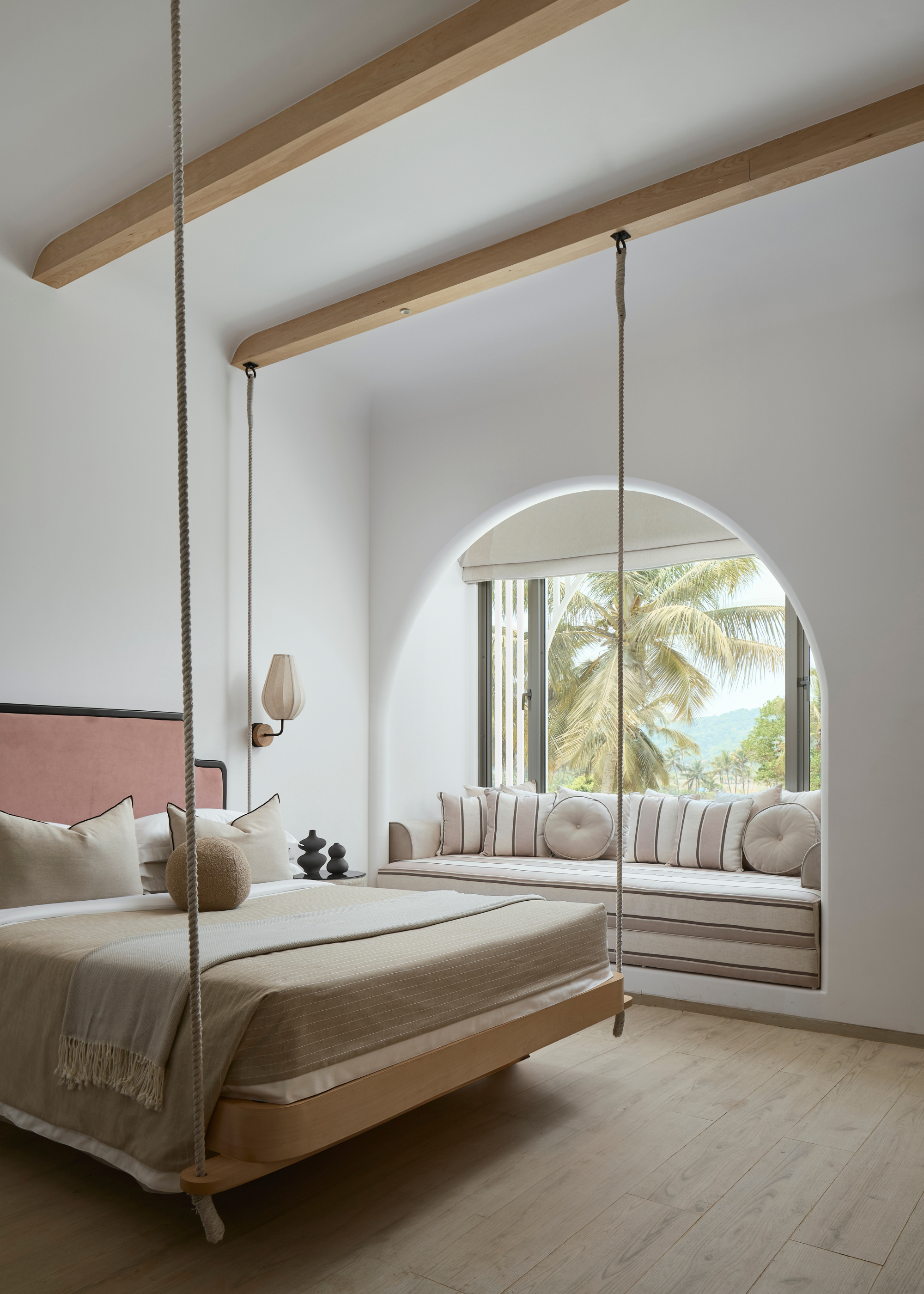 A bedroom with wood beams on the ceiling supporting a hanging bed, a wall sconce by the bedside, and a cozy nook with pillows by an arched opening near the window