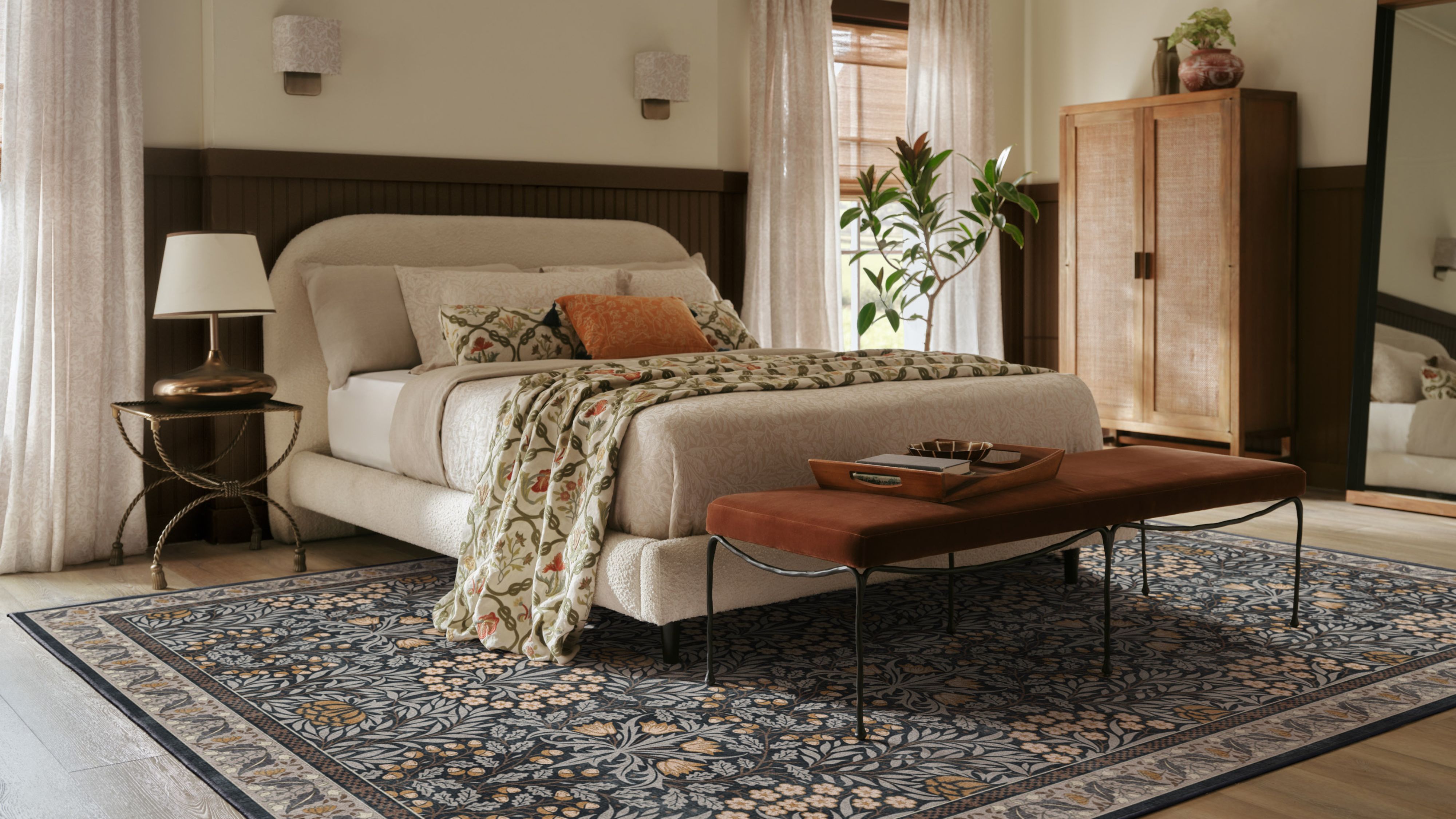 Warm, richly layered bedroom featuring a sculptural cream bed frame, terracotta iron ottomans, and a moody floral rug.