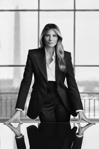 Black and white photo of FLOTUS Melania Trump standing at desk in front of window with Washington Monument in background