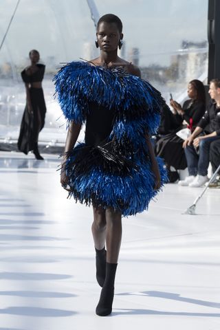 Catwalk with a focus on a model wearing a navy blue / black midi length frill / tinsel dress and black ankle boots.