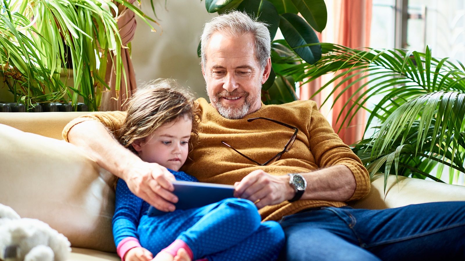 A grandfather and granddaughter sit on a couch using a digital device.