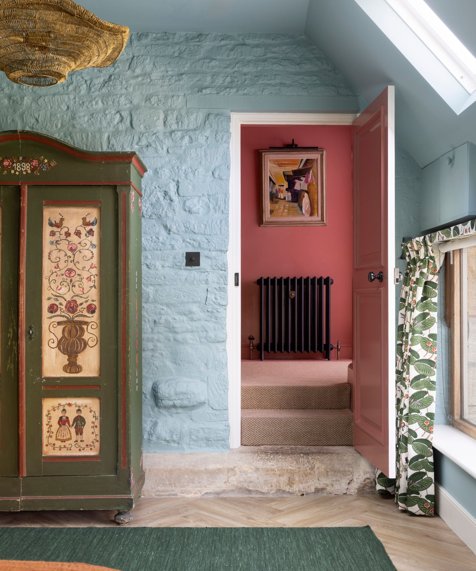 A bedroom with a brick wall painted powder blue, a doorway leading into a pink room, and a vintage green wardrobe.