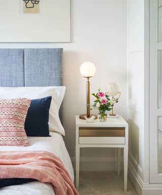 A white bedroom with a light blue bed, white bedding with a pink and dark blue throw pillow, a pink throw blanket, and a white nightstand with white and purple flowers and a brushed brass lamp