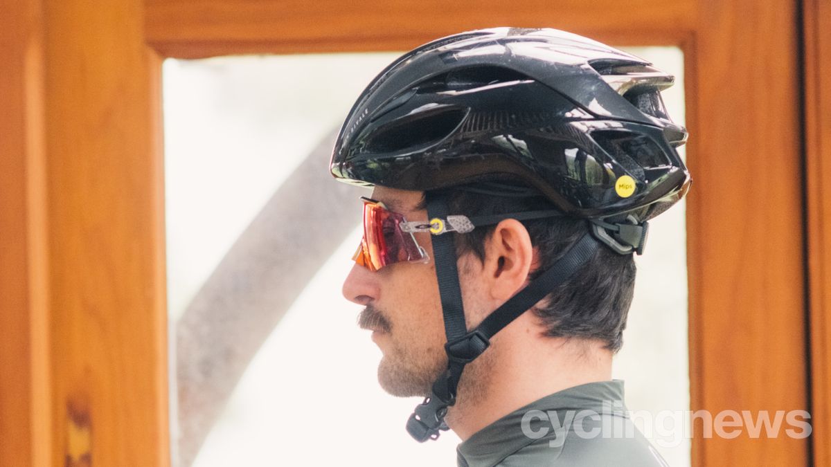 A white man with a moustache wears a red pair of Oakley Kato glasses under a black helmet