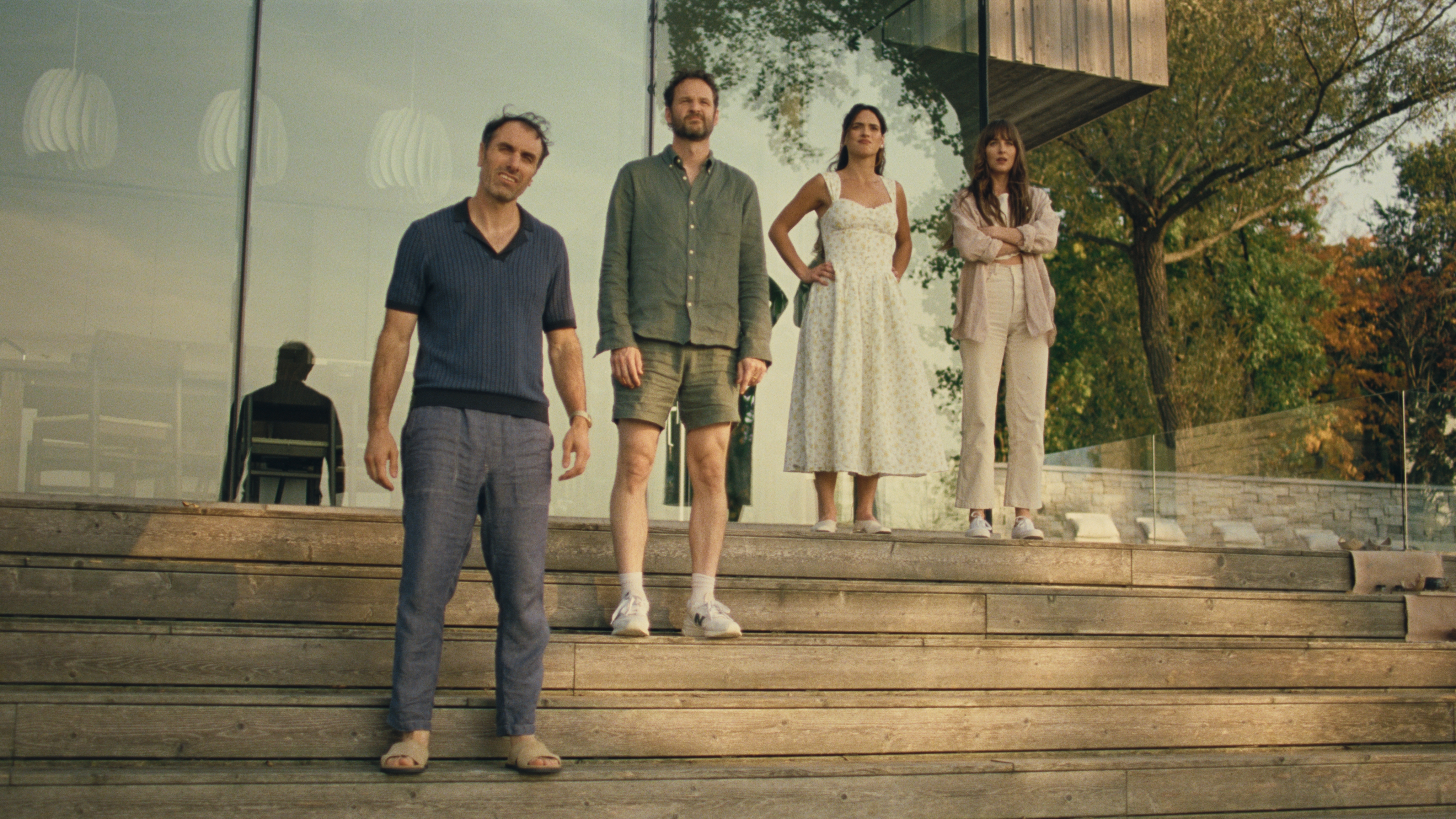 Michael Angelo Covino, Kyle Marvin, Adria Arjona, and Dakota Johnson stand at the top of steps outside of a home looking into the distance in a still from the movie SPLITSVILLE