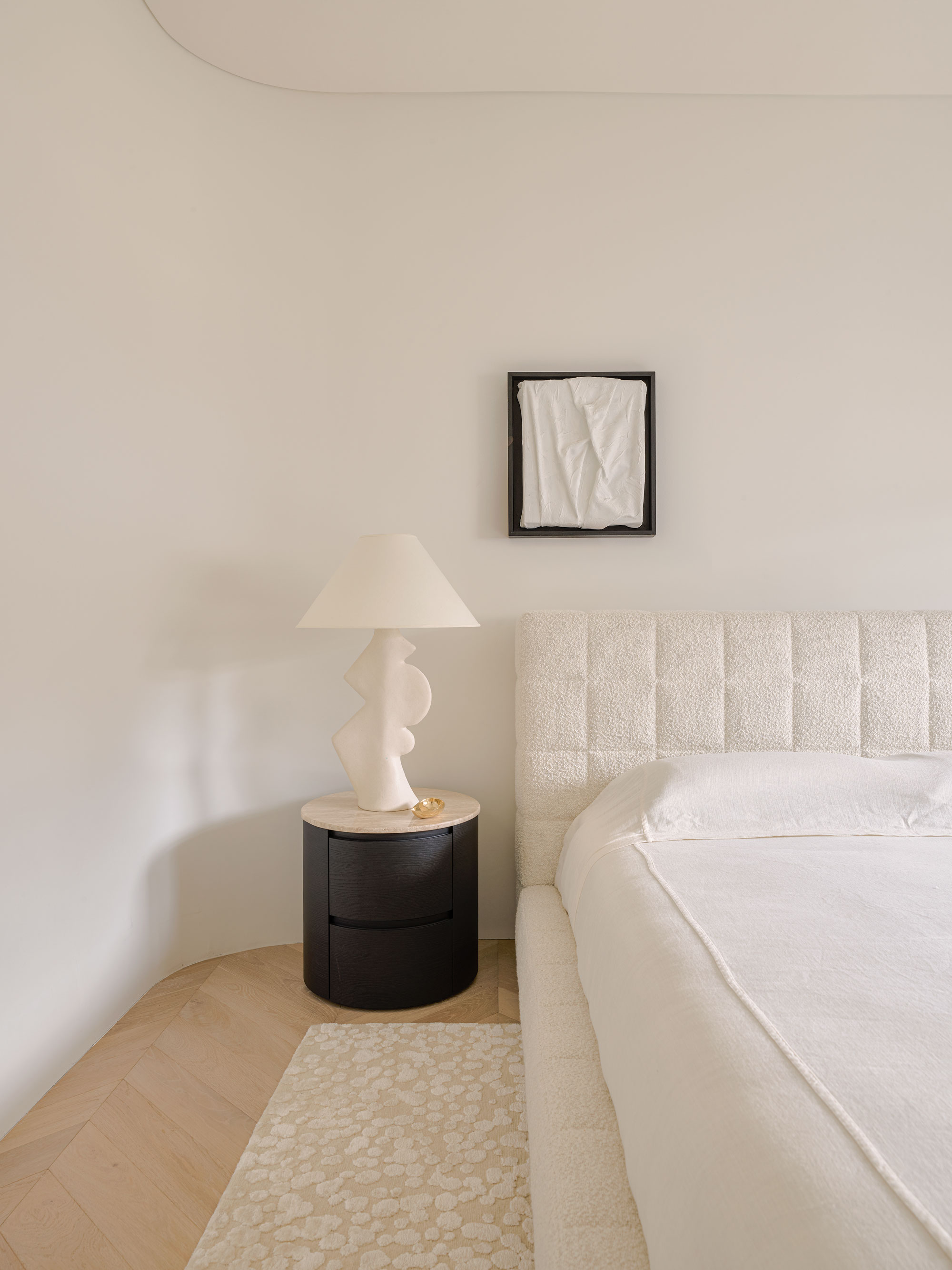 Cream colour drenched bedroom, the bed has a fabric frame and headboard, with a black and wood nightstand, a white sculptural lamp and an art work.