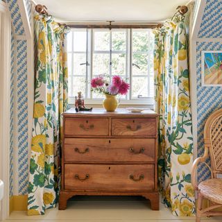 yellow and green floral curtains around bay window which houses a wooden chest of drawers in room with blue striped wallpaper