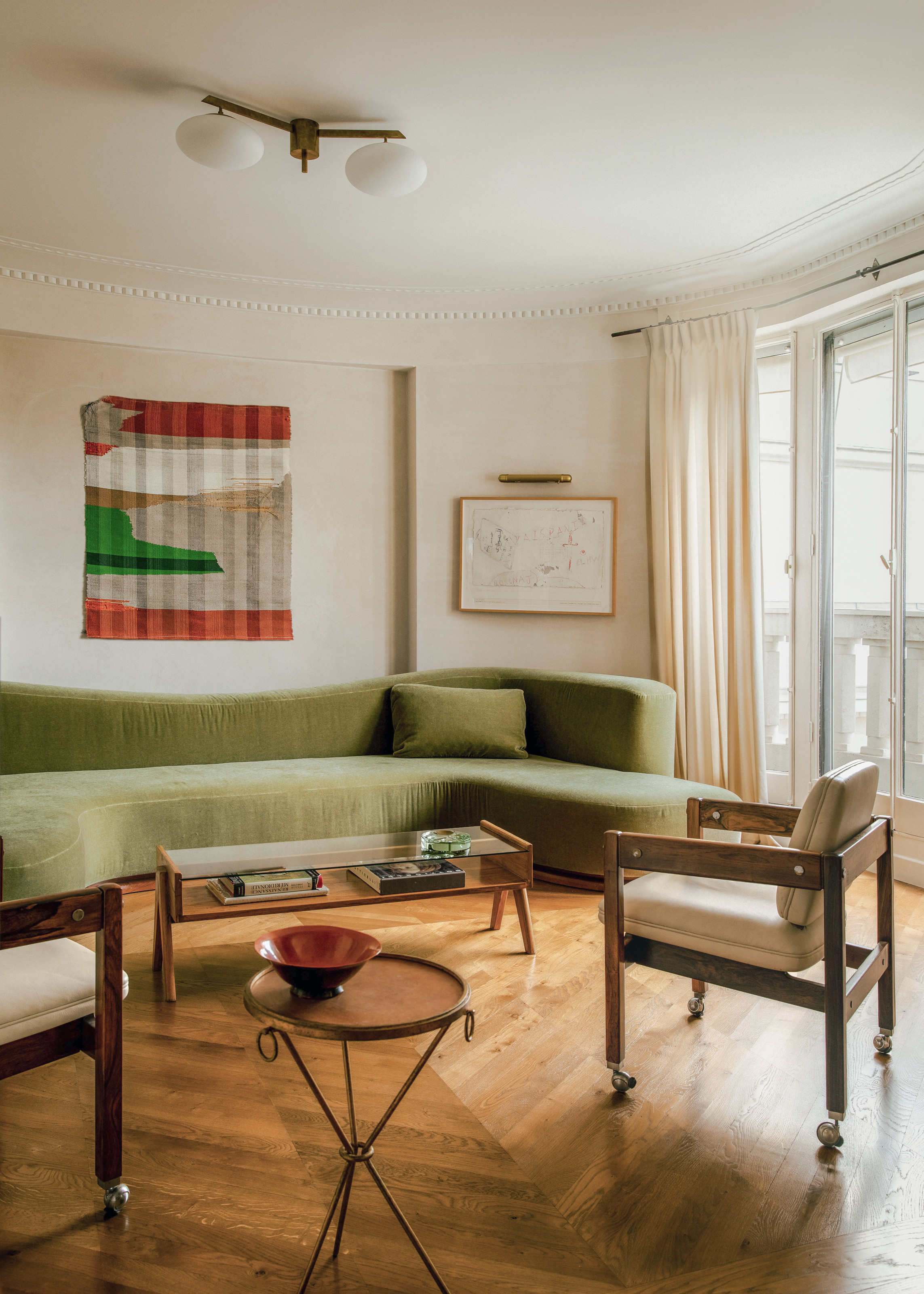 Living room with green velvet curved sofa, glass and wood coffee table, vintage wooden chairs on castor wheels and elegant metal side table
