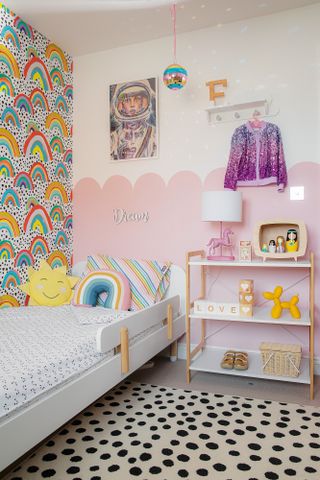 Kid's room with rainbow feature wallpaper on one wall, pink scalloped paint effect on the other. White bed with rainbow and sun throw cushions, black and white spotty rug, Scandi open shelving and astronaut print over the bed