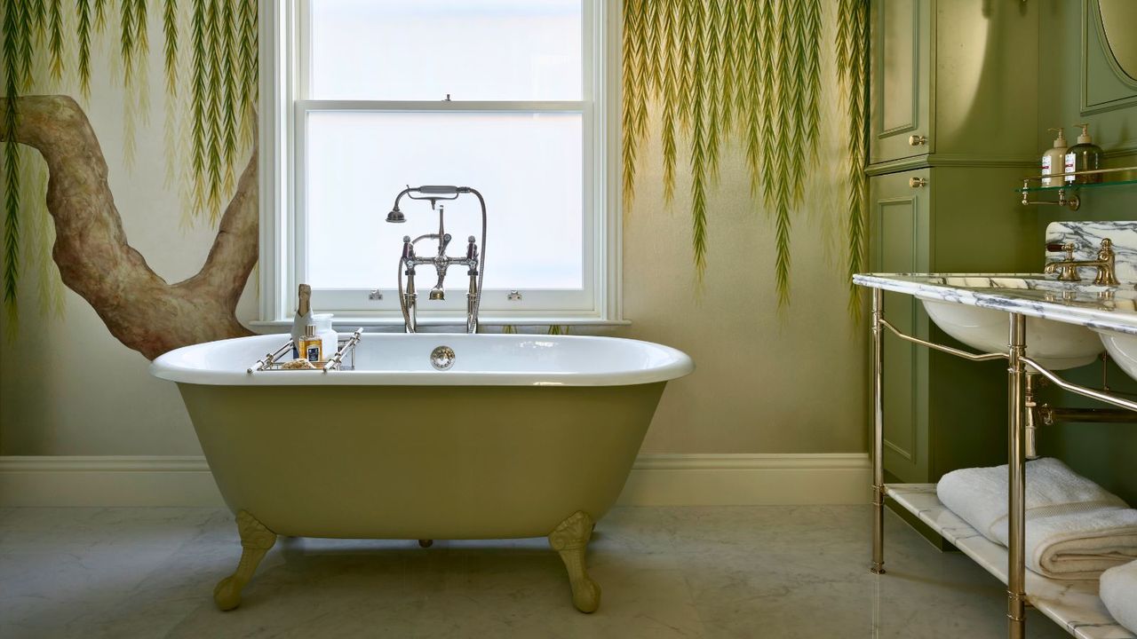 Large green bathroom with freestanding bath and large window to the back. There is a weeping willow tree wallpaper across the back wall