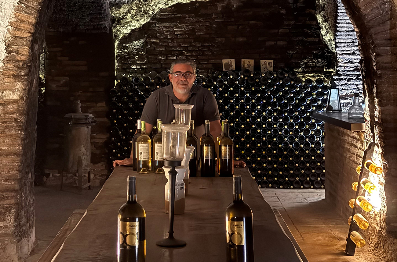 Albert Ustrell Serrano, standing in a dark cellar full of wines, behind a large table with bottles of wine on it
