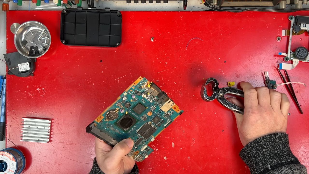 Left Hand holding PS2 motherboard with pliers in right hand over red workbench.