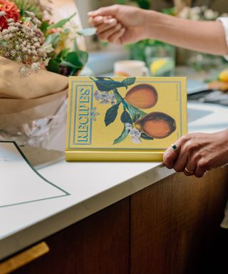 image of recipe book on white countertop being held, wrapped flowers by the sink