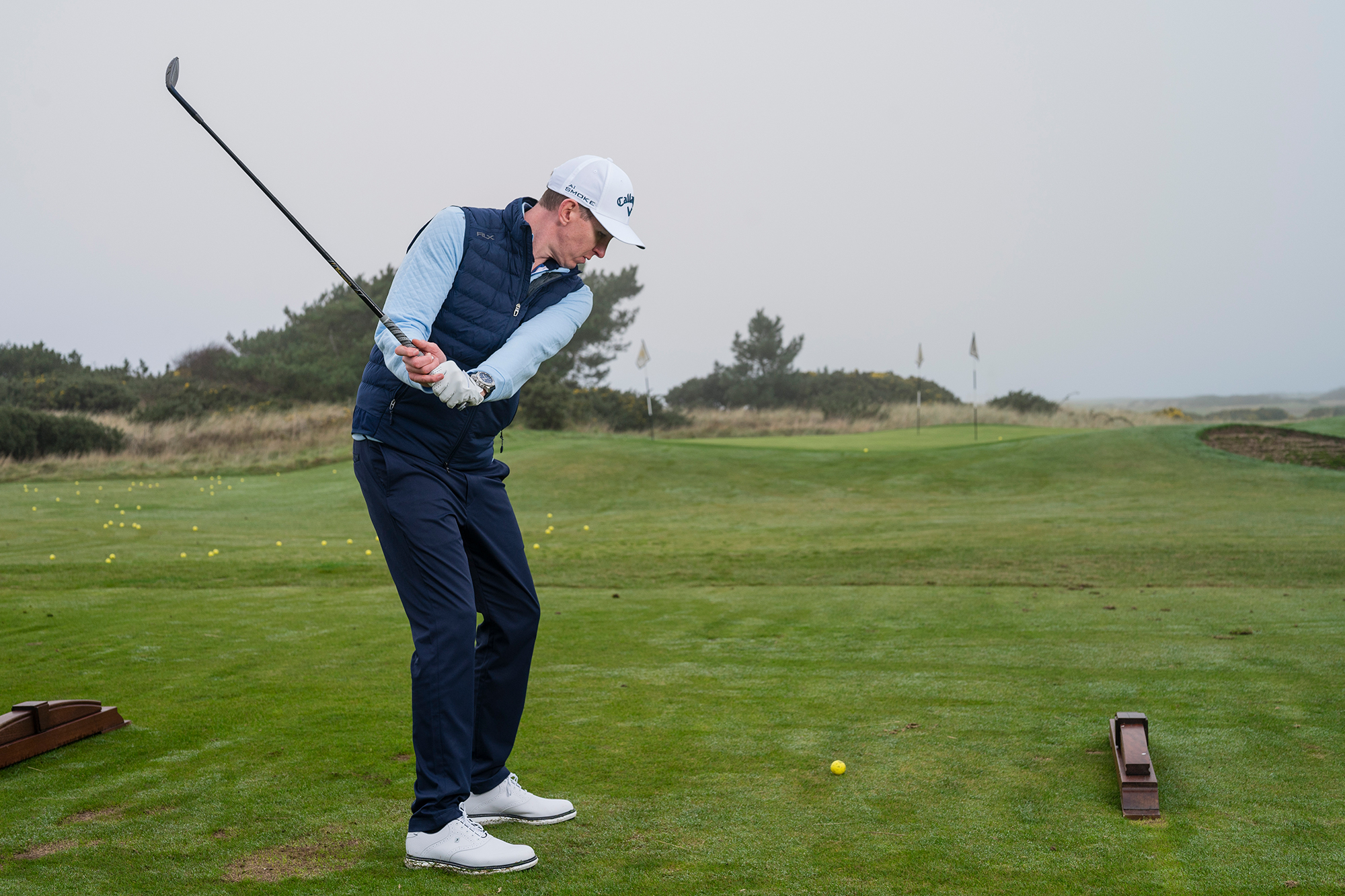 Ben Emerson at the top of his backswing with a wedge, on the short game practice area at the driving range of Trump Turnberry, with two golf balls already on the green giving him feedback on the distance control