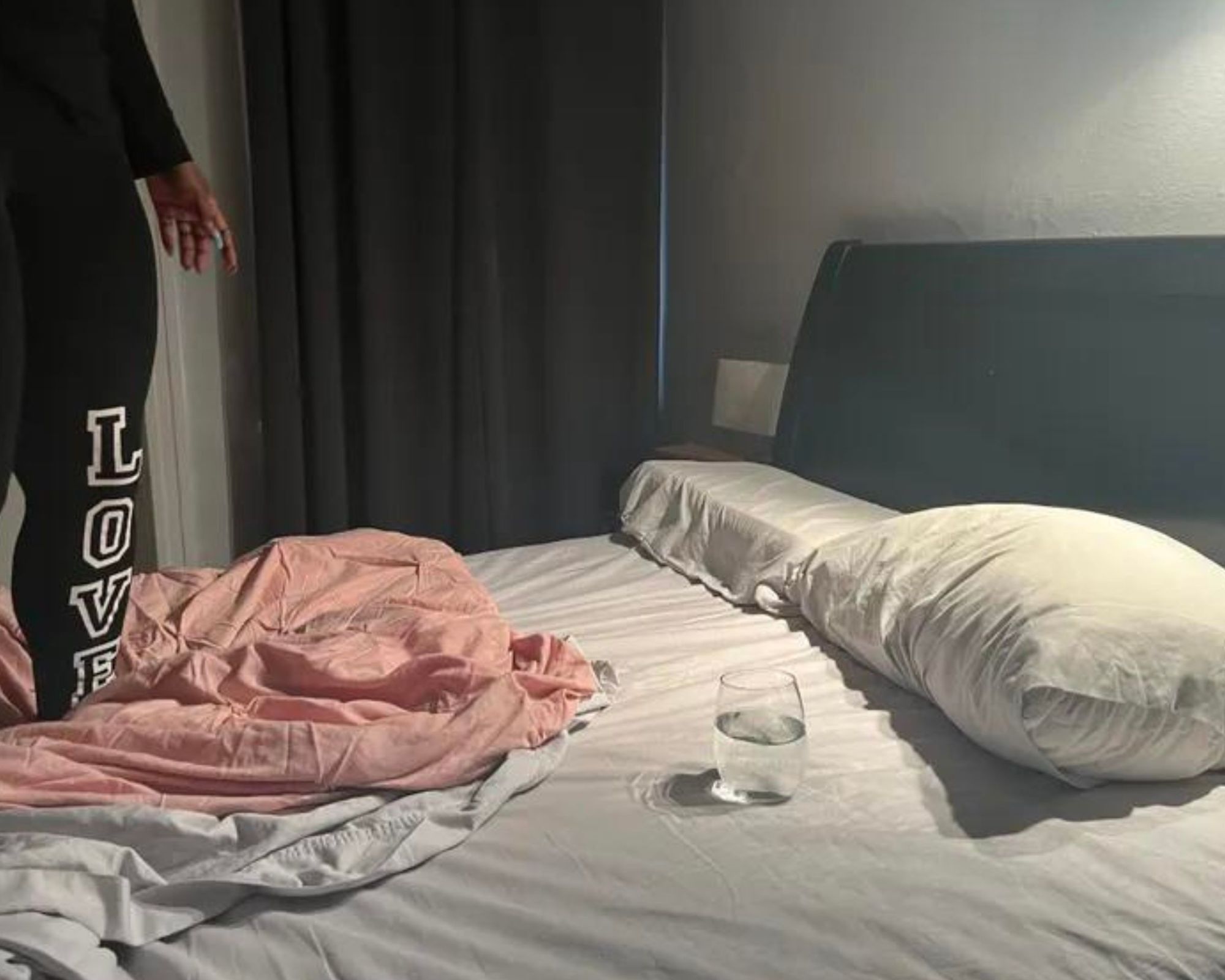 A woman wearing black leggings with 'Love' spelt out in white lettering standing to the left of the image on the Siena Memory Foam Mattress, with white sheets, pillows in white pillowcases, and two bunched up sheets in pink and white at her feet. Also on the bed is full a glass of water. The headboard is black, matching the black curtain in the background of the image, and the walls are white.