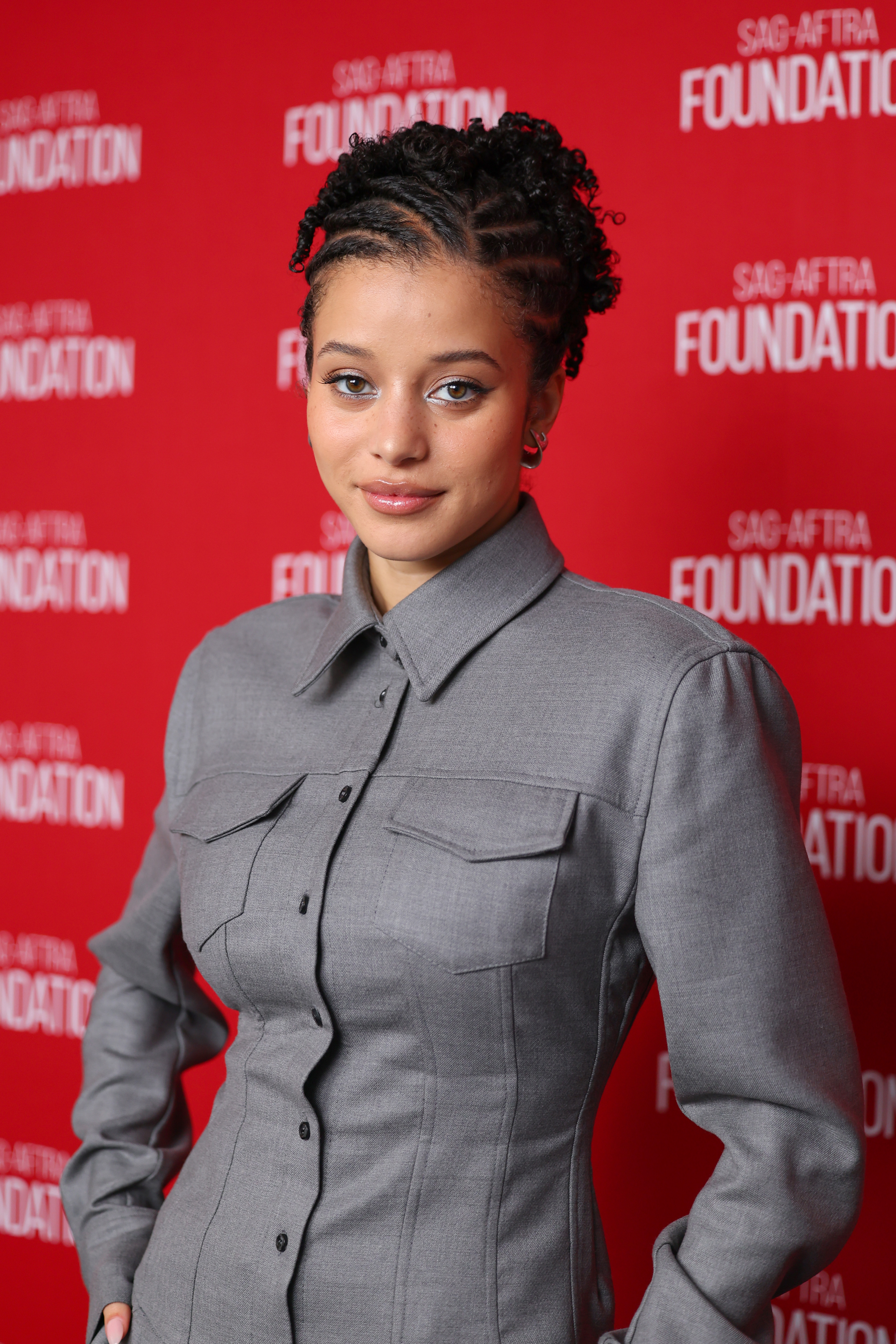 a young woman poses in front of a sag-aftra backdrop wearing a gray button down dress