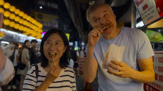 phil rosenthal from somebody feed phil walking through an asian night market and eating food on a stick