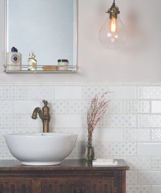 A bathroom with a silver mirror with a shelf, white subway tiles with a blue pattern, a dark wooden vanity with a white basin and a hanging pendant light
