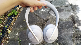 En hand håller upp ett par vita Bose Noise Cancelling Headphones 700 över en asfalterad gångväg.