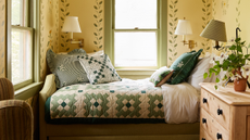 a bedroom nook with a patchwork bedspread, several pillows, a wooden dresser, wall sconces, and yellow wallpaper with ivy motifs