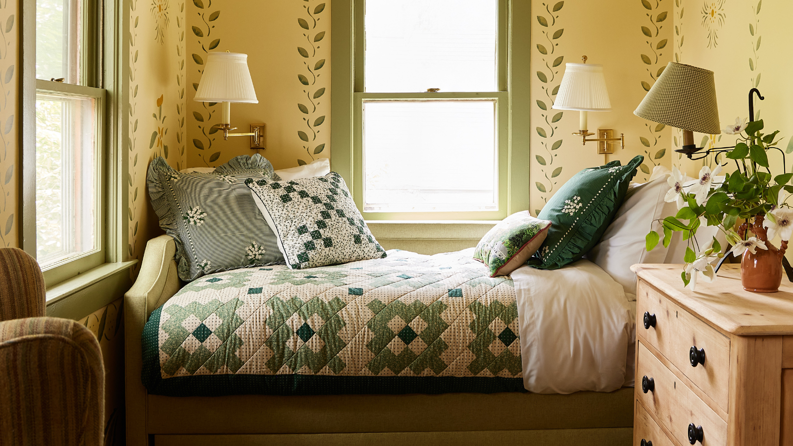 a bedroom nook with a patchwork bedspread, several pillows, a wooden dresser, wall sconces, and yellow wallpaper with ivy motifs