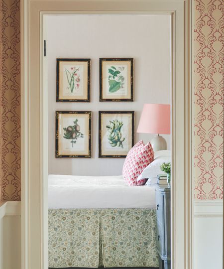 A white doorway with pink and white wallpaper either side and white baseboards showing a view into a bedroom, with a bed with white bedding, a green floral bedskirt, and a pink and white cushion. The walls are pale pink, and there is a floral gallery wall. Next to the bed is a blue bedside table in the foreground, and white ceramic lamp with pink lampshade in the background.